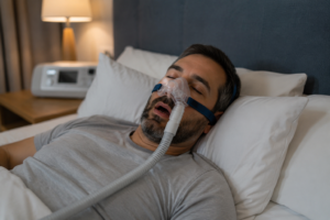A middle-aged man sleeping peacefully in a softly lit bedroom while wearing a CPAP mask connected to a sleep apnea machine, illustrating home treatment for sleep apnea.